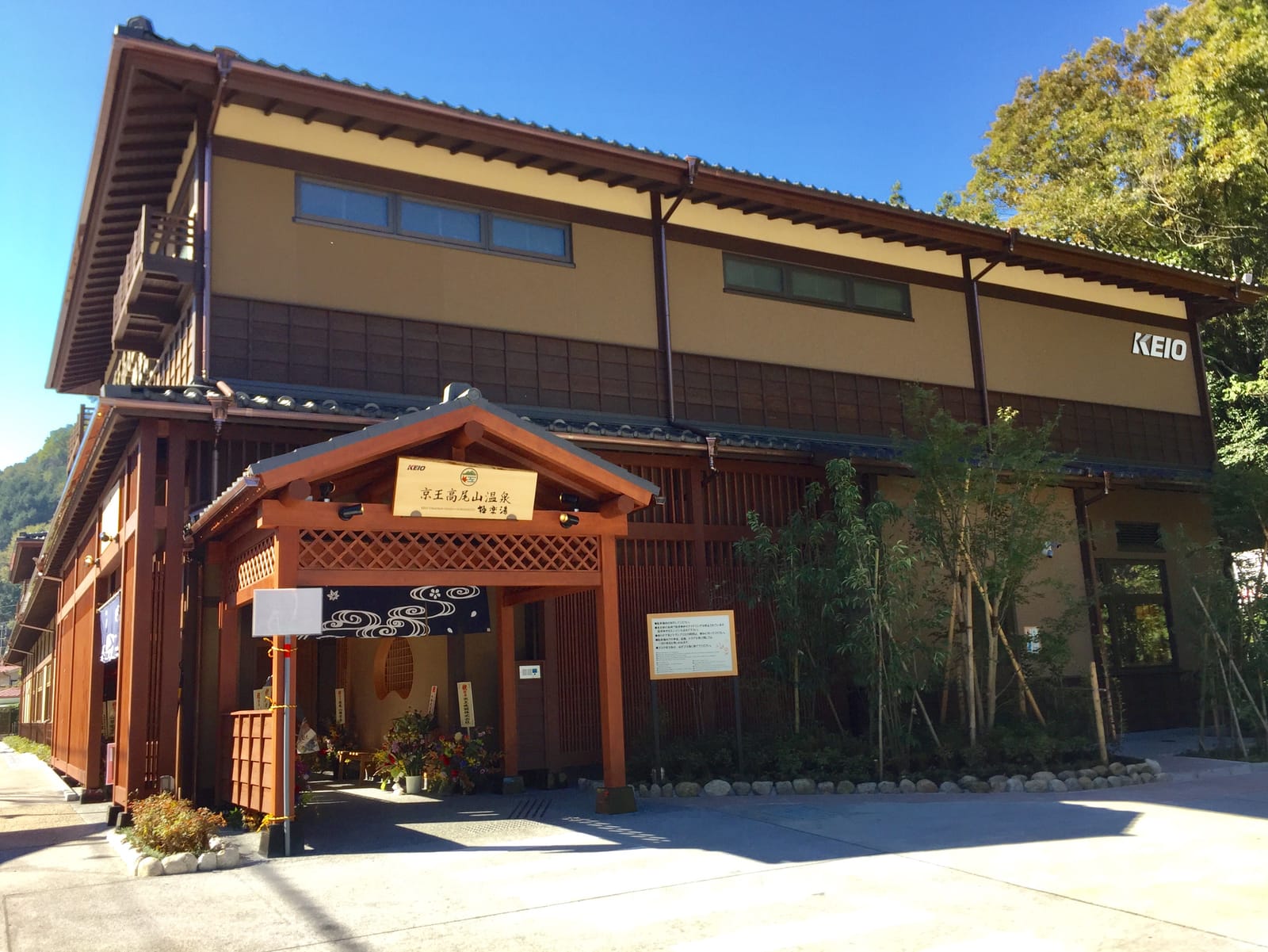 The wooden entrance and building of Keio Takaosan Onsen Gokurakuyu, with autumn trees behind.