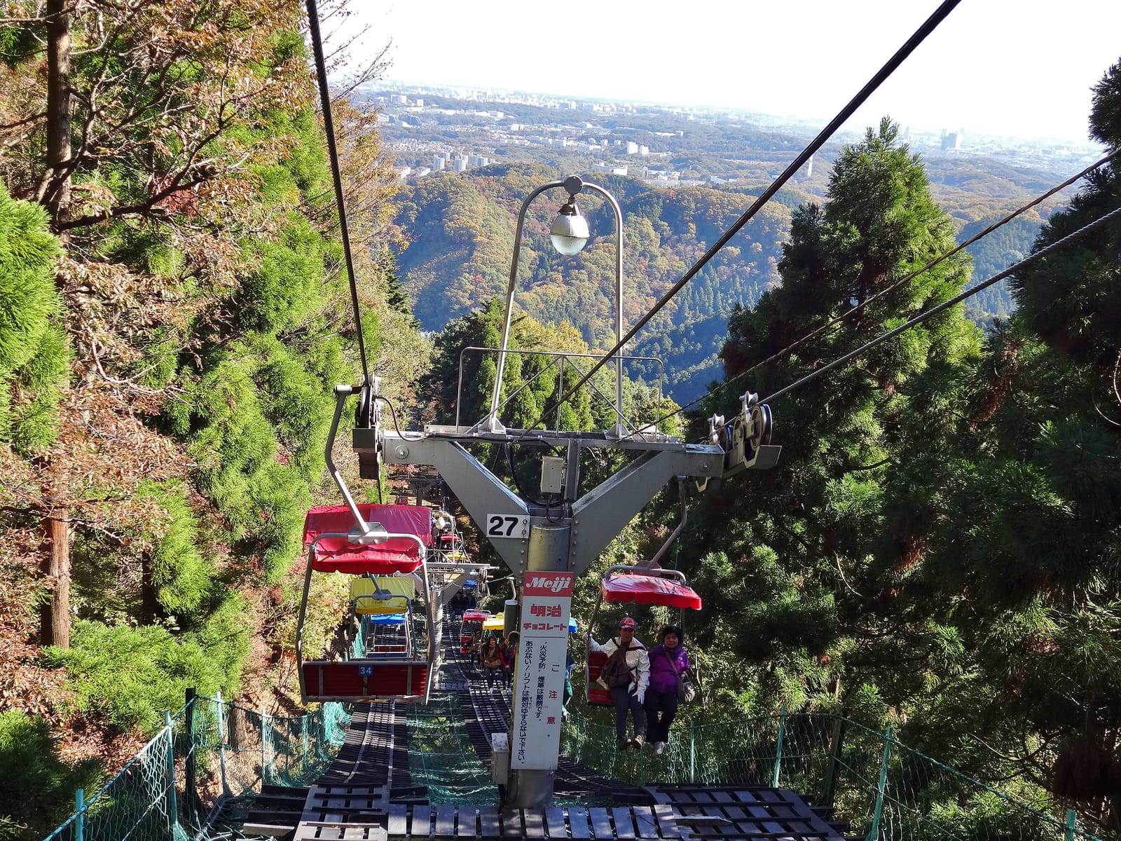 Riders on the Mt. Takao two-seat chairlift climbing through cedars in autumn, with the plain in the distance.